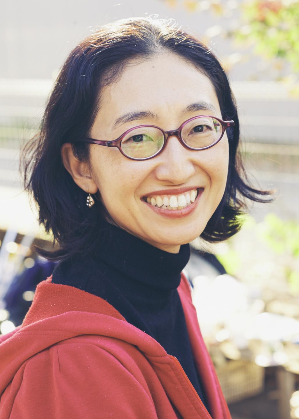 Photo of Sawako Nakayasu by Mitsuo Okamoto. Sawako, a Japanese woman with medium-short black hair of uneven length, is wearing a red hoodie, black turtleneck, one visible earring, and purple glasses. She is slightly angled, looking this way with a big smile. The background is outdoors and mostly blurry.